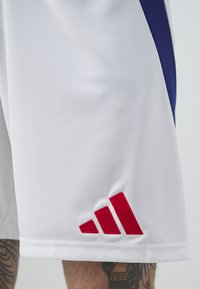 Pantalones cortos deportivos blancos, confeccionados con una tela suave, que cuentan con un panel lateral azul y un logotipo bordado de Adidas en rojo en el dobladillo inferior.