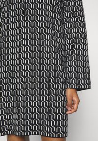 Robe à manches longues à motif géométrique noir et blanc, montrant le bras et la main droite sur un fond clair uni.