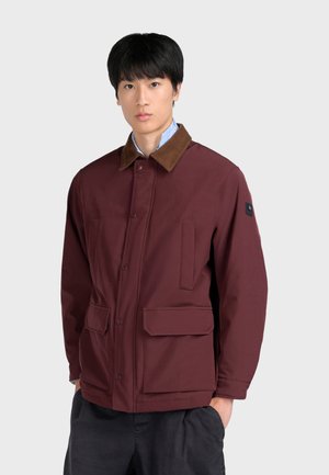 Jeune homme aux cheveux noirs porte une veste bordeaux avec un col marron et des poches à rabat, debout devant un fond gris clair uni.