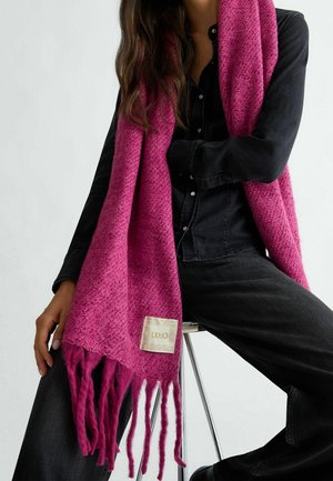 Femme assise sur un tabouret, vêtue d'une tenue en denim noir et d'une longue écharpe texturée magenta avec des franges et une étiquette visible.