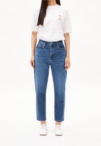 Džínsy s vysokým pasem v modré barvě se rovnými nohavicemi, obsahující pět kapes a mírným vyblednutím, v kombinaci s bílým tričkem s logem.