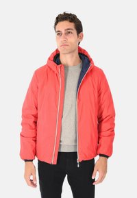 Uomo con una giacca con cappuccio rossa sopra un maglione grigio e pantaloni neri, in piedi davanti a uno sfondo chiaro e semplice.