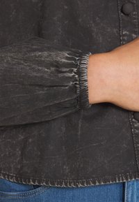 Czarna, teksturowana bluzka z marszczonymi mankietami i detalami z guzikami, o lekko błyszczącym, przetartym wyglądzie. Miękka tkanina o luźnym kroju.