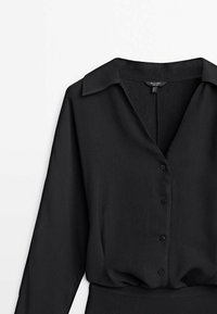 Blouse noire à manches longues avec col, boutonnage sur le devant et tissu légèrement texturé, présentée sur fond blanc.