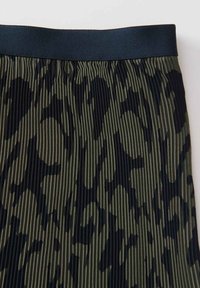Jupe verte plissée avec un motif camouflage noir, dotée d'une ceinture élastique noire. Le tissu texturé ajoute de la profondeur au design.