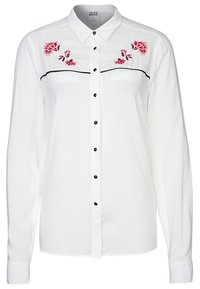 Bílá halenka s dlouhým rukávem, černými knoflíky, předními kapsami a květinovou výšivkou v růžové a červené barvě na ramenou, s klasickým límečkem.