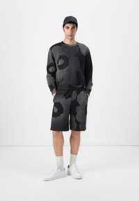 Sweatshirt- und Shorts-Set in Schwarz und Grau mit Muster, aus einem Stoff mit weicher Textur. Weiße Sneakers und helle Socken runden das Outfit ab.
