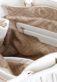 Valkoinen nahkainen käsilaukku, jonka sisällä on vaaleanbeige kangaspäällyste, varustettuna vetoketjullisella taskulla ja toistuvalla kultapainatettu logokuvio.