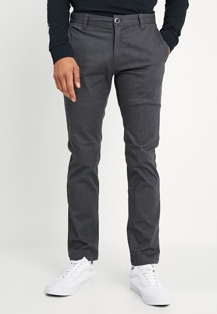 Homme portant un pantalon gris foncé slim, des baskets blanches et une chemise noire à manches longues, debout avec une main dans la poche.