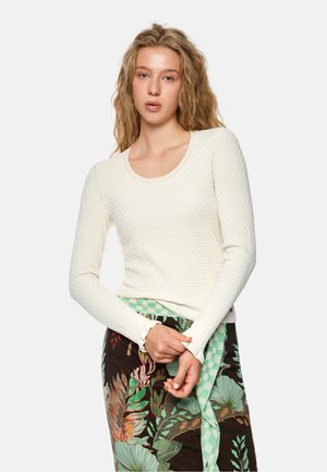 Cremfarbene, strukturierte Langarmbluse mit rundem Ausschnitt. Kombiniert mit einem gemusterten Rock, der tropische Blätter und geometrische Designs zeigt.