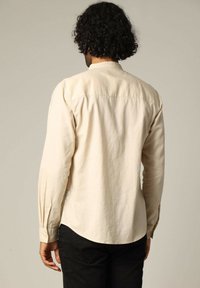 Beige langemouwen overhemd met een button-down kraag, gestructureerde stof, schouderpas en manchetten. Draag met zwarte broek, vanuit de achterkant gezien.