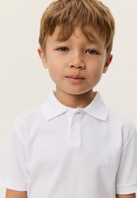 Λευκό πόλο πουκάμισο με κλασικό γιακά, διαθέτει δύο κουμπιά και μικρό κεντημένο λογότυπο στο στήθος. Λείος βαμβακερός υφασμάτινος ιστός.