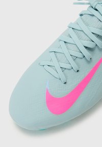 Chaussure de football bleu clair en matériau texturé avec un swoosh Nike rose. Elle présente des lacets fins et plats ainsi qu'un design de orteil arrondi.
