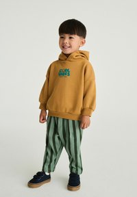 Brun hætte sweatshirt med "COOL VIBES" grafik, parret med grønne stribede bukser og mørke sneakers. Glat tekstur, afslappet pasform.