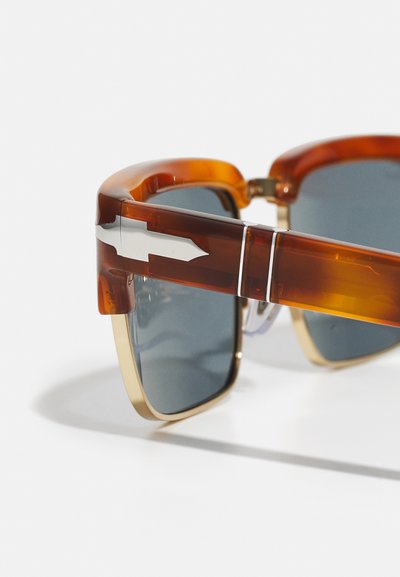 Óculos de sol em padrão tartaruga com uma armação de plástico espessa, lentes cinzentas e detalhe metálico em forma de seta nas hastes. Destaques em dourado acentuam o design.