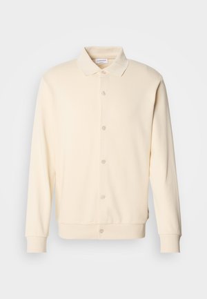 Hanorac cu butoni de culoare crem, cu mâneci lungi, guler cu rever și manșete ribbordate. Textură netedă, design minimalist.