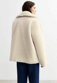 Krämfärgad plyschjacka med bred krage och texturerad tyg. Löst sittande med sänkta axlar, tillsammans med mörkblå denimbyxor.