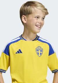 Gul fotbollströja med blå accenter, med Sveriges nationella emblem, tre randiga axlar och texturerad blommönster.