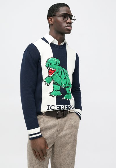Άνδρας που φορά γυαλιά, μπλε σκούρο και λευκό πουλόβερ με πράσινο δεινόσαυρο και το κείμενο "ICEBERG", μπεζ παντελόνια και σκούρη ζώνη, μπροστά σε απλό φόντο.