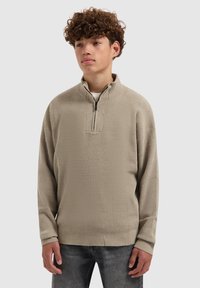 Beige kwart-rits pullover gemaakt van textuurstof, met een hoge kraag en lange mouwen. Draag het over een wit shirt en combineer het met donkere spijkerbroeken.