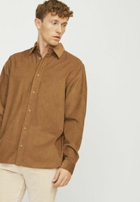 Chemise marron à boutons, en tissu texturé, avec un col pointu et des manches longues, associée à un pantalon en velours côtelé de couleur claire.
