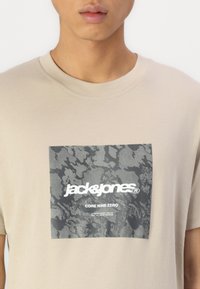 Camiseta de algodón beige con un diseño gráfico gris que presenta el texto "jack & jones" y "CORE NINE ZERO" en blanco. Cuello redondo clásico.