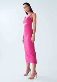 Robe cintrée rose avec détail découpé, fines bretelles et ourlet mi-long. Associée à des sandales dorées à talons fins et lanières. Texture lisse.