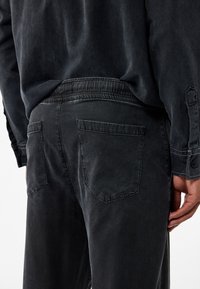 Schwarze Denim-Hosen mit strukturierter Oberfläche, die eine lockere Passform, einen elastischen Bund und zwei Gesäßtaschen mit kantigen Klappen aufweisen.