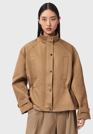 Nainen, joka pukeutunut vaaleanruskeaan korkeakauluksiseen takkiin, jossa on mustat napit, läppätaskut ja leveät hihat, yhdistettynä vastaaviin vaaleanruskeisiin plisseehousuihin.