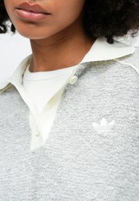 Gaiši pelēks, teksturēts džemperis ar baltu apkakli un izšūtu Adidas logo; ar pogu plāksni un slāņotu baltu iekšējo kreklu.