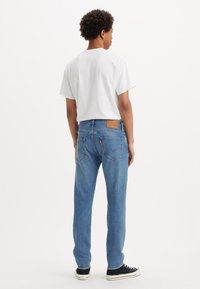 Jeans en denim bleu clair avec une coupe droite, patch en cuir à l'arrière et deux poches arrière, portés avec un t-shirt blanc uni et des baskets noires.