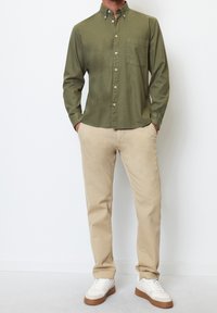 Olivově zelená košile na knoflíky s dlouhými rukávy a náprsní kapsou, spárovaná s béžovými kalhotami a bílými teniskami, na jednobarevném pozadí.