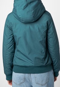 Veste à capuche teal avec une texture lisse, ourlet côtelé et coutures visibles au dos. Portée avec un jean bleu clair.
