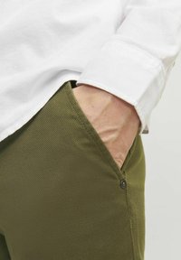 Olive green woven pants featuring a side pocket with a black button, paired with a white collared shirt. Textured fabric visible.