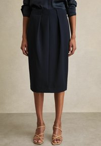 Tumši zila midi svārki ar krokām, piegrieztā dizainā, gluda tekstūra. Valkāti kopā ar smilškrāsas augstpapēžu sandālēm ar siksniņām.