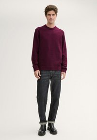 Pull en laine bordeaux avec un col rond et des poignets côtelés, associé à un jean foncé et des bottines noires, présenté sur un fond uni.