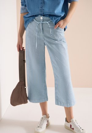Personne portant un pantalon large à rayures bleues et blanches, une chemise en denim, des baskets blanches, tenant un sac bandoulière marron par la sangle.