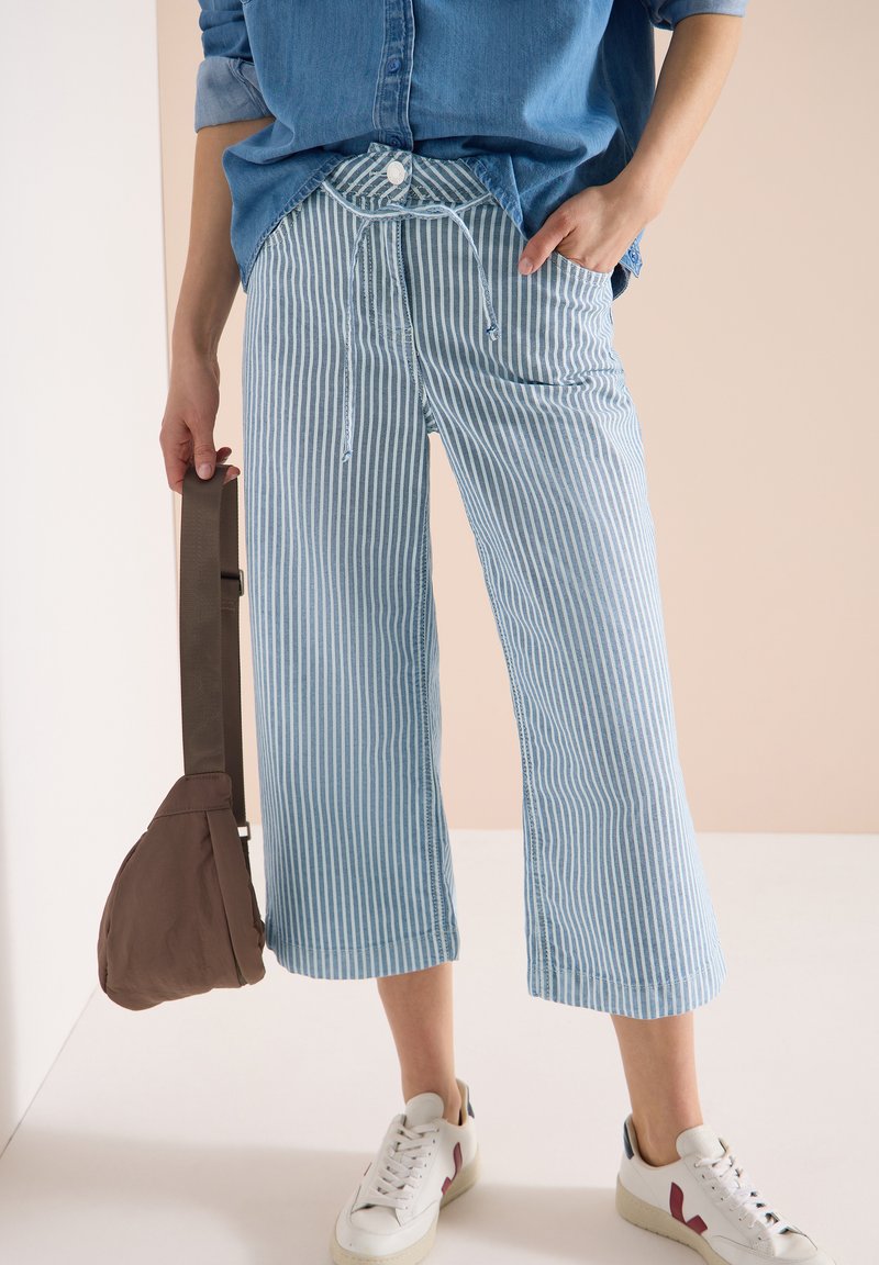 Personne portant un pantalon large à rayures bleues et blanches, une chemise en denim, des baskets blanches, tenant un sac bandoulière marron par la sangle.