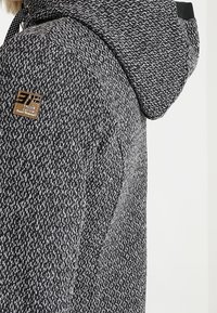 Hættetrøje lavet af tekstureret grå og sort stof, med et lille emblem på ærmet og en kontrastfyldt indvendig foring.