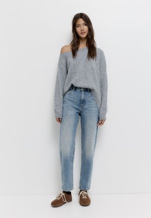 Kvinde iført en løs grå off-shoulder sweater, lyseblå højtaljede jeans og brune snørebåndssko, stående mod en hvid væg.