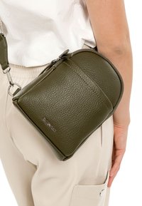 Tuvplāns ar tumši zaļu teksturētu plecu somu, kas nēsāta ap vidukli virs bēšām biksēm un baltas krekla, redzams sudraba rāvējslēdzējs un zīmola logotips.