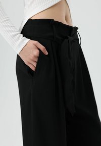 Zwarte hooggetailleerde broek met een strikceintuur en zijzakken, met een gladde textuur en een ontspannen pasvorm.