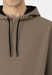 Primo piano di una persona che indossa una felpa con cappuccio color taupe con coulisse nere e fodera interna del cappuccio nera.