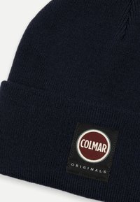 Sötét tengerészkék kötött sapka bordás textúrával és hajtogatott mandzsettával. Egy kerek fekete címke található rajta, fehér és piros "COLMAR" és "ORIGINALS" felirattal.