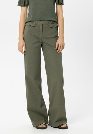 Frau trägt olivgrüne Hose mit weitem Bein und aufgesetzten Taschen mit Knöpfen sowie ein passendes kurzärmeliges Oberteil, steht vor einem weißen Hintergrund.