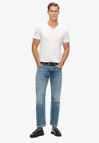 Biała koszulka z dekoltem w serek, niebieskie jeansy z wybledzonym wykończeniem, czarny pasek i czarne mokasyny. Model stoi z rękoma w kieszeniach.