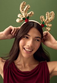 Gold sequin reindeer antlers on a fabric headband, adorned with green leaves and a red pom-pom, worn by a person with long brown hair.