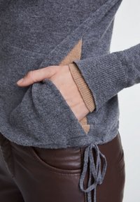 Grijze gebreide trui met een brede zak die een contrasterende beige voering toont. Voorzien van geribbelde manchetten en een trekkoord onderrand.