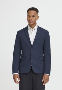 Sötétkék zakó texturált anyagból, két gombbal, kifordított kollárral és két elülső zsebbel, világos csíkos ing felett viselve.