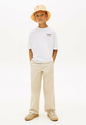 Chłopiec w brzoskwiniowym kapeluszu typu bucket, białej oversize koszulce z logo, beżowych spodniach o szerokich nogawkach i beżowych sneakersach, stojący z rękami w kieszeniach.